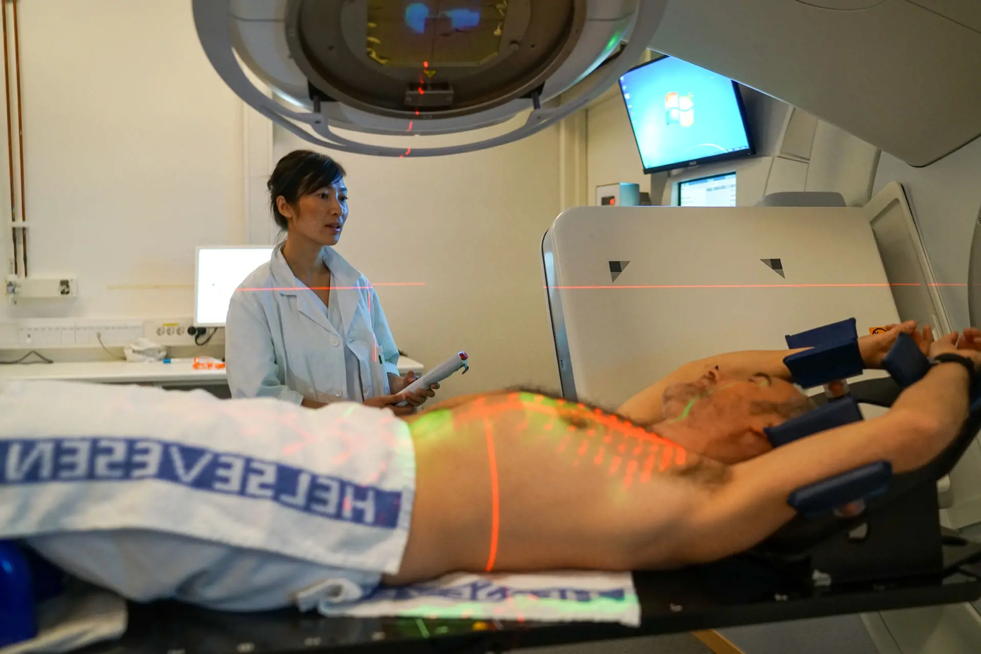Mann som liggger på en behandlingsbenk med en strålemaskin over seg. Strålemerker vises på mannens bryst og mage. Lege ser på.
