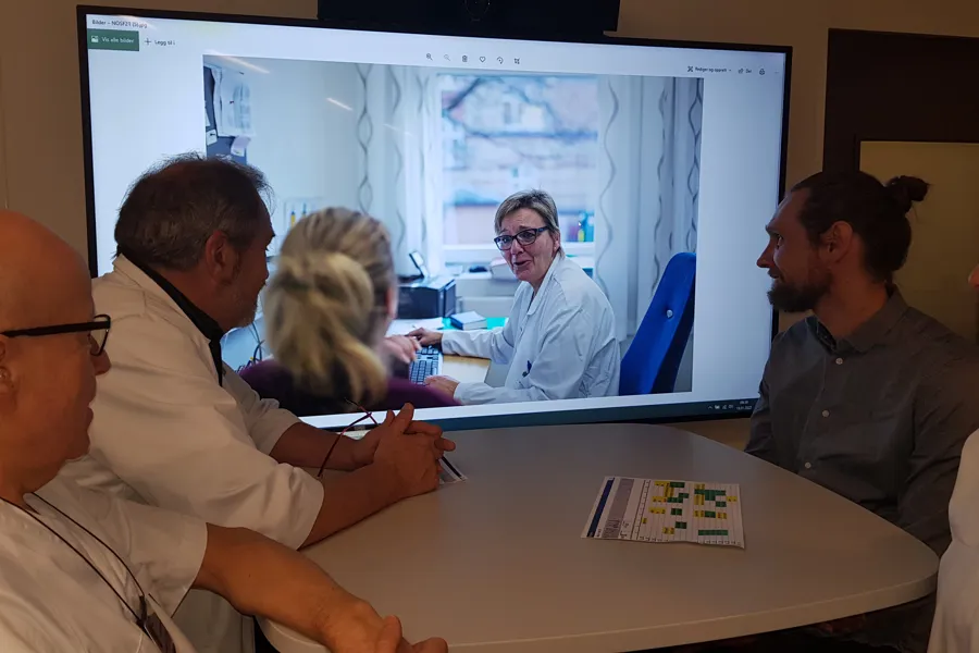4 leger som sitter og ser på en videoskjerm. På videoskjermen ser de en lege som snakker med en pasient. Videokonsultasjon.