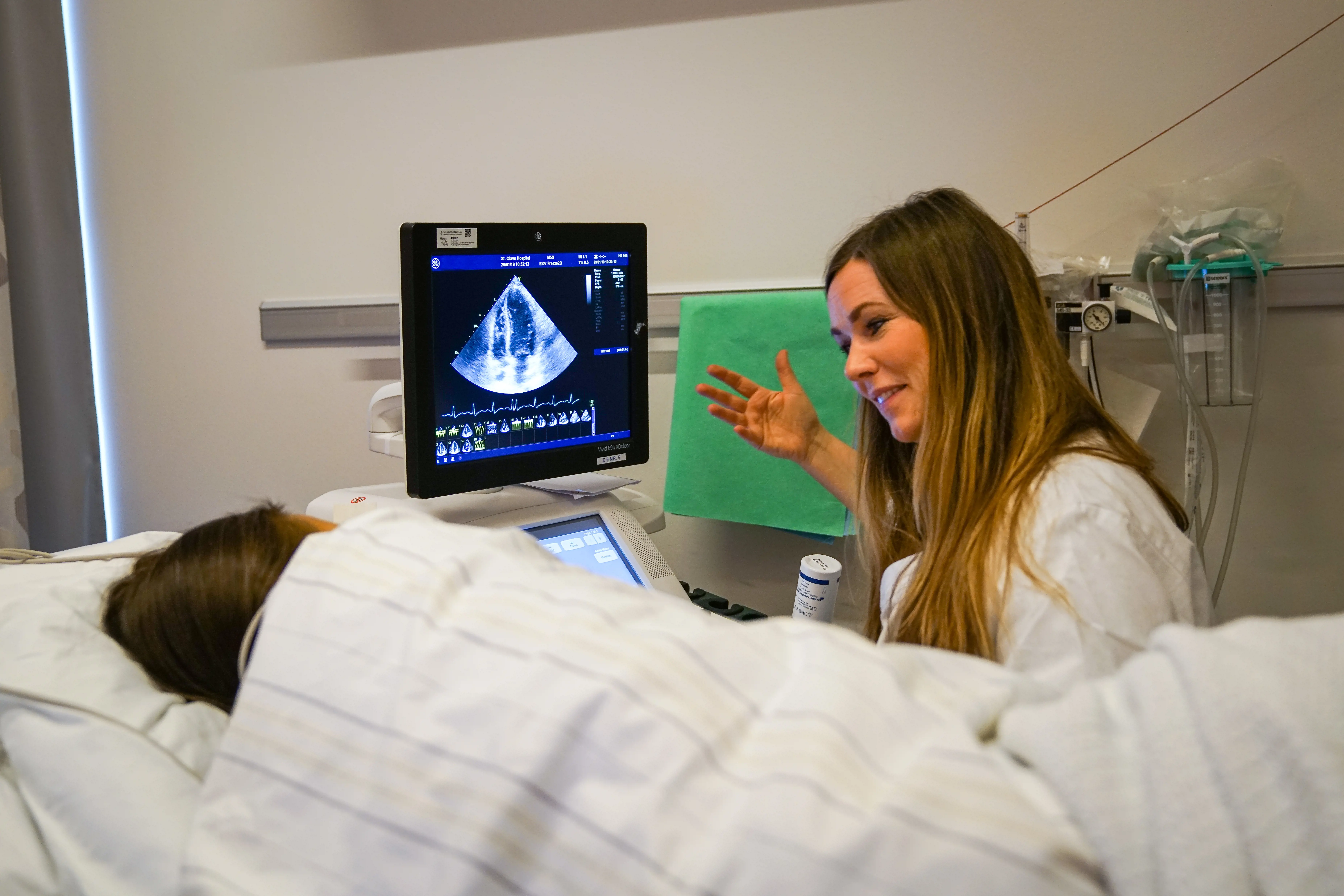 En lege som viser en pasient et ultralydbilde på en pc-skjerm. Pasienten ligger i sengen, under dyna med ryggen til kameraet.