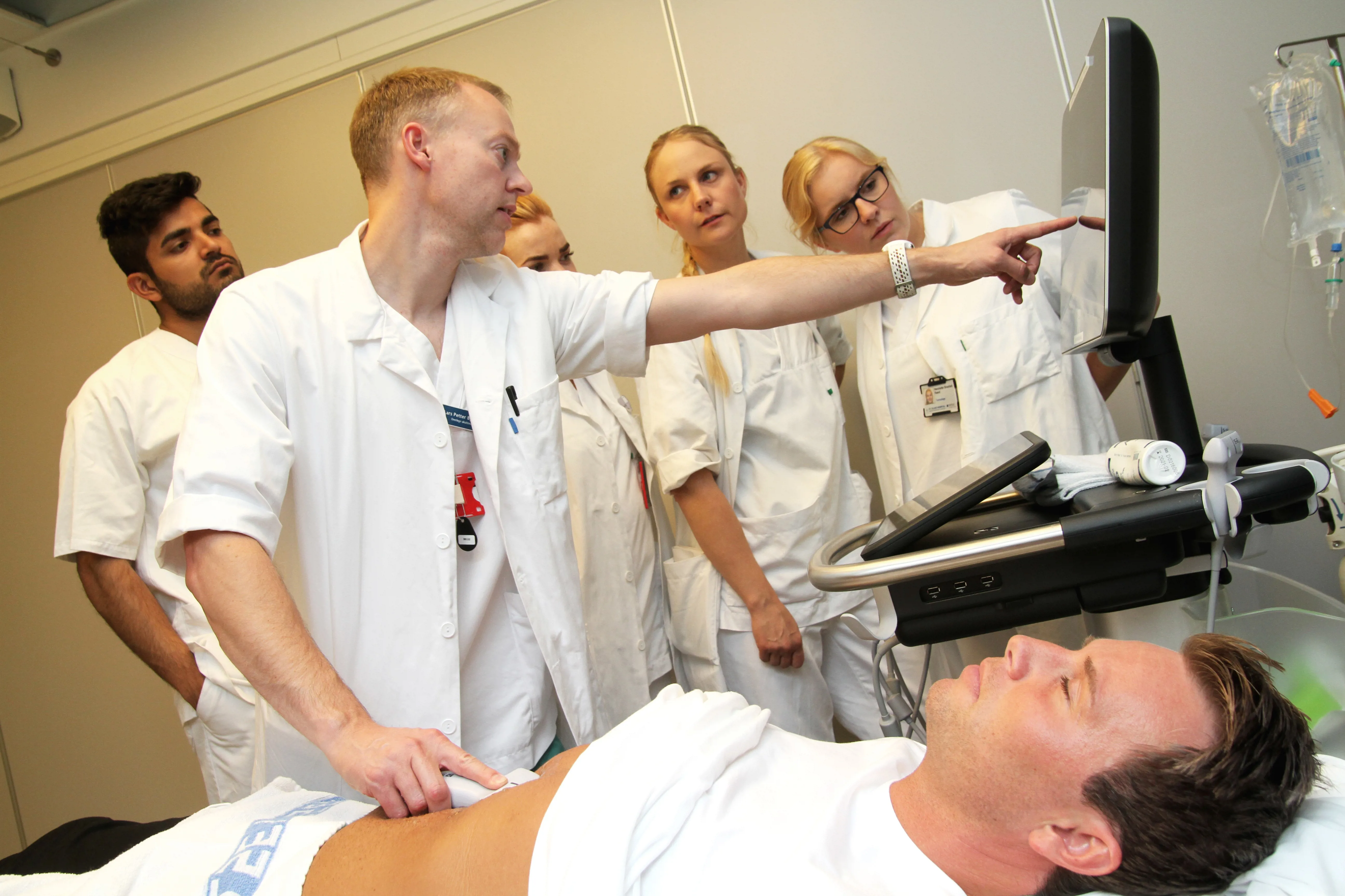 En lege holder en ultralydprobemot magen til en pasient som ligger på ryggen på en seng. Legen peker på en skjerm. 