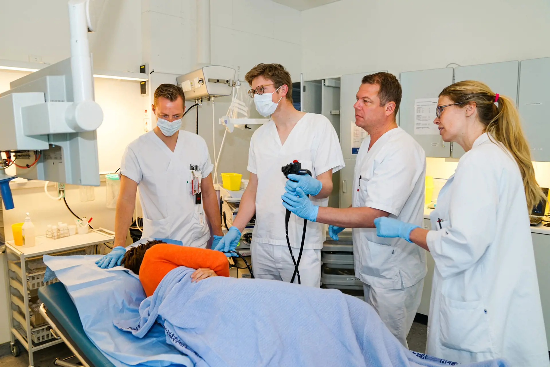 En LIS-lege blir rettledet av en erfaren lege når han skal utføre gastroskopi. To medleger ser på. En pasient ligger på sengen.